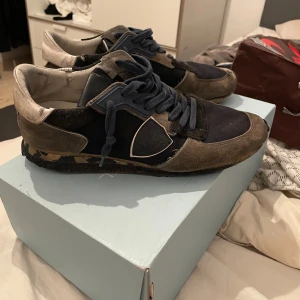 Philippe Model sneakers i blått och grått - Säljer ett par Philippe Model sneakers i blått och grått. De har en cool, sliten look med snörning och en bekväm passform. Perfekta för en avslappnad stil. Skorna är i bra skick och redo för nya äventyr!