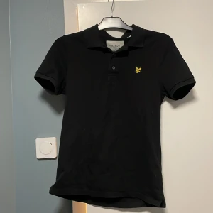 Svart pikétröja från Lyle & Scott - Säljer en snygg svart pikétröja från Lyle & Scott i storlek S. Tröjan är i bra skick och har en klassisk design med korta ärmar och en liten broderad logga på bröstet. Perfekt för både vardag och fest! 