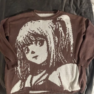 Brun stickad tröja med anime-motiv - Säljer en unik brun stickad tröja med ett stort anime-motiv i beige. Tröjan har långa ärmar och ribbstickade muddar vid ärmslut och nederkant. Perfekt för höst och vinter, och för alla anime-fans där ute! 🧡