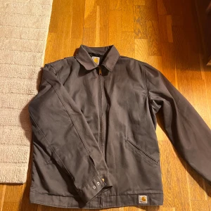 Grå jacka från Carhartt - Säljer en grå Carhartt-jacka i mycket bra skick. Den har en dragkedja framtill och är quiltad på insidan för extra värme. Perfekt för höst och vår! Jackan har långa ärmar och två praktiska fickor. En klassisk och stilren design som funkar till det mesta.
