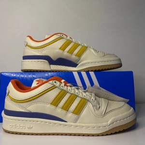 Säljer ett par Adidas Forum Low Wood Wood sneakers i vitt med gula och blå detaljer. Skorna har en snygg retrostil med orange insida och klassisk snörning. Perfekta för både vardag och fest! Kommer i originalkartong.