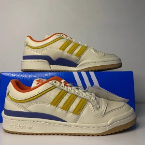 Adidas Forum Low Wood Wood - Säljer ett par Adidas Forum Low Wood Wood sneakers i vitt med gula och blå detaljer. Skorna har en snygg retrostil med orange insida och klassisk snörning. Perfekta för både vardag och fest! Kommer i originalkartong.