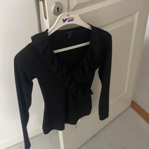 Svart långärmad topp med volang - Säljer en elegant svart topp  i storlek S. Den har långa ärmar och en snygg volangdetalj runt halsringningen. Perfekt för både vardag och fest! 🖤