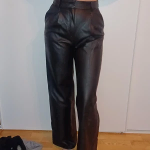 Svarta skinnbyxor - Säljer ett par snygga svarta skinnbyxor i mycket bra skick. De är högmidjade och har en straight passform som ger en cool look. Perfekta för höst och vinter, och funkar både till vardags och fest. Byxorna har en dragkedja och knappar framtill. De är fodrade på insidan.