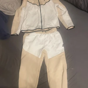 Beige och vit träningsdräkt från Nike - Säljer en snygg och bekväm träningsdräkt från Nike i beige och vitt. Setet består av en jacka med dragkedja och matchande byxor. Perfekt för träning eller en avslappnad dag. Jackan har svarta detaljer och byxorna har en elastisk midja för bästa passform.