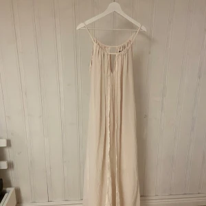 Champagnefärgad långklänning Bik Bok - Elegant champagnefärgad långklänning med en luftig och flödande design. Klänningen har en enkel och stilren look med smala axelband och en lätt rynkad detalj vid halsen. Perfekt för sommarens fester eller en avslappnad dag på stranden.