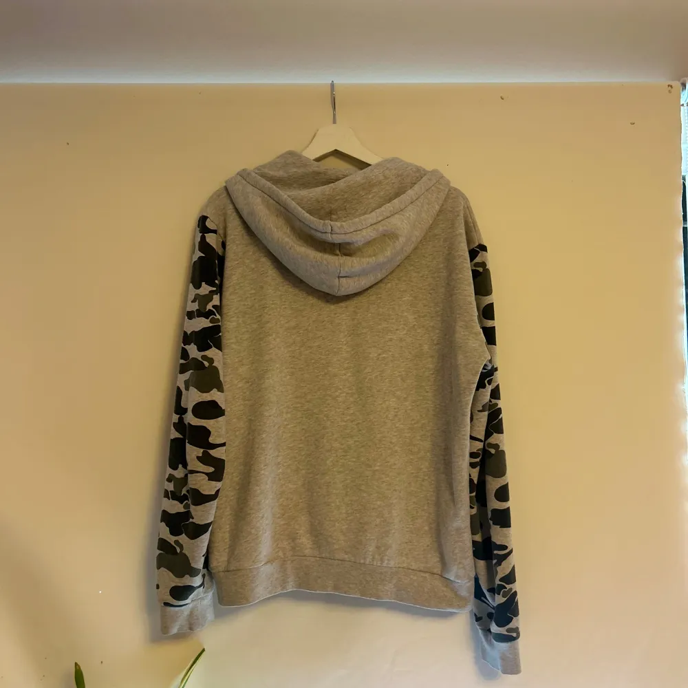 Säljer en snygg grå hoodie med coola camouflageärmar och ficka. Perfekt för en avslappnad stil och passar bra till både höst och vår. Den har en justerbar huva med snören och är i mycket bra skick. Perfekt för dig som gillar streetwear!. Tröjor & Koftor.