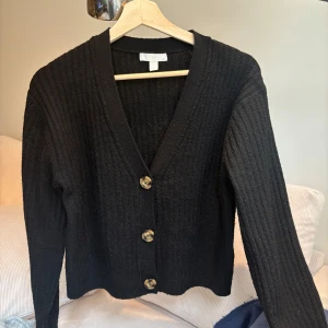 Svart ribbad kofta med knappar - Säljer en svart ribbad kofta med stora knappar framtill. Den har en v-ringad design och långa ärmar, perfekt för höst och vinter. Koftan är i mycket bra skick!