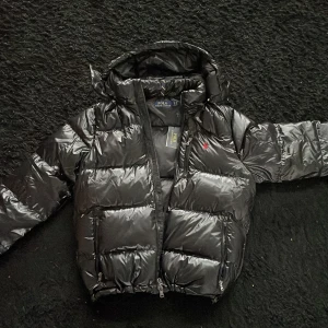Svart dunjacka från Polo Ralph Lauren - Säljer en snygg svart dunjacka från Polo Ralph Lauren. Jackan är i mycket bra skick och har en glansig finish med dragkedja framtill. Den har långa ärmar och en praktisk huva. Perfekt för kalla vinterdagar! 🖤