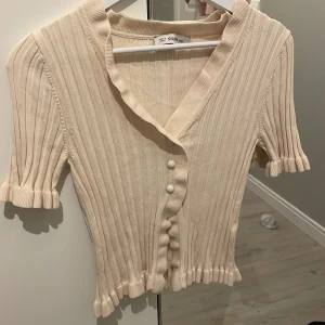 Beige ribbad kofta från Dilvin - Säljer en superfin beige ribbad kofta från Dilvin. Den har korta ärmar och knappar framtill. Perfekt för både vår och sommar! Koftan är i ett mjukt och bekvämt material, perfekt att ha över en topp eller klänning.