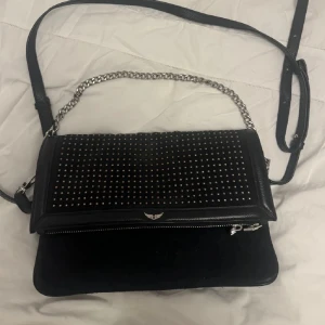 Svart axelväska  - Snygg svart axelväska med silverfärgade nitar och kedjedetaljer. Väskan har en justerbar axelrem och dragkedja för säker förvaring. Perfekt för både vardag och fest!