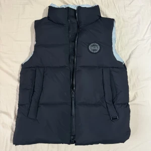 Svart Canada Goose väst (Black Label) - Säljer en svart dunväst från Canada Goose i mycket bra skick. Västen har en dragkedja framtill och ett stilrent märke på bröstet. Perfekt för kyliga höst- och vinterdagar. Den är både varm och trendig, idealisk för lager-på-lager-stil. Storleken är M men passar som M-L.