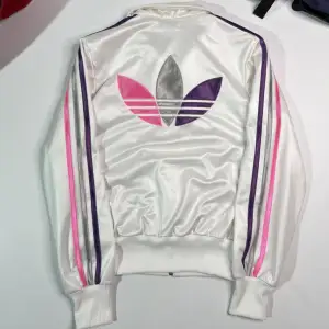 Säljer en snygg vit Adidas jacka i glansigt material med rosa och lila ränder längs ärmarna. Adidas-logga i rosa på bröstet och stor logga på ryggen.