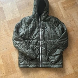 Grön quiltad jacka från Nike - Säljer en grön quiltad jacka från Nike i storlek M. Jackan har en dragkedja framtill och en huva för extra skydd. Perfekt för höst och vår med sin stilrena design och bekväma passform. Nike-loggan är snyggt placerad på bröstet. 🧥