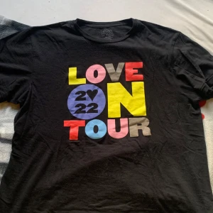 Svart Harry Styles t-shirt - Säljer en svart Harry Styles t-shirt i bomull med färgglatt tryck 'Love On Tour 2022' på framsidan. Baksidan har en lista över turnéstäder i gult. Perfekt för fans! 💛En liten målarfläck på baksidan se bild