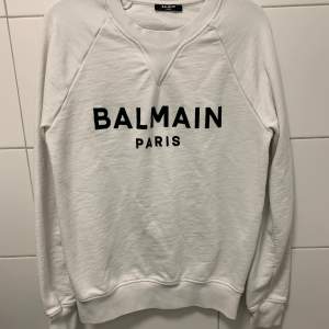 Säljer en snygg vit sweatshirt från Balmain med svart text på bröstet. Tröjan är i mycket bra skick och perfekt för både vardag och fest. Den har långa ärmar och en klassisk passform. Superbekväm och stilren! 🖤