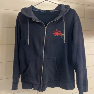 Vintage 90s stüssy  - Säljer en mycket sällsynt mörkblå hoodie från Stüssy med dragkedja och huva. Den har en cool logga på framsidan och grön på baksidan. Perfekt för en avslappnad stil. Lite slitage på muddarna, men annars i bra skick. Perfekt för kallare väder! Storlek M men passar som S/Xs. Skriv vid minsta funderingar!
