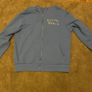 Blå hoodie med rosdetalj - Säljer en snygg blå hoodie från Porsche Design. Den har en dragkedja framtill och en stor rosdetalj i strass på baksidan. Perfekt för en avslappnad stil med lite extra bling. Passar både till vardags och festliga tillfällen! Liten fläk på magen