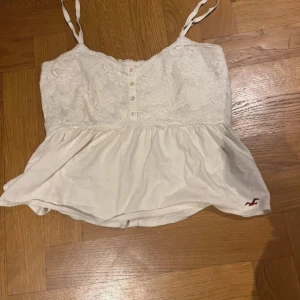 Vit spetslinne från Hollister - Säljer ett superfint vitt linne från Hollister i storlek XS. Toppen har spetsdetaljer och knappar framtill, vilket ger en romantisk touch. Perfekt för sommardagar eller en avslappnad festkväll. Den är i mycket bra skick och redo för nya äventyr!