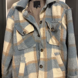 Rutig teddyjacka i blått och beige - Säljer en supermysig teddyjacka i rutig design med blå, beige och vita toner. Jackan har långa ärmar och knappar framtill. Perfekt för höst och vinter när du vill hålla dig varm och stilren. Två praktiska fickor med lock på bröstet. Passar både till vardags och lite mer avslappnade tillfällen.
