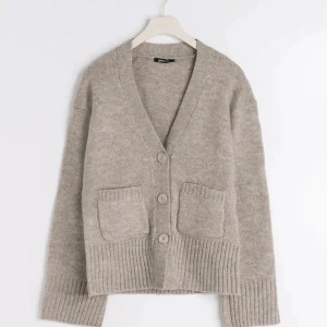 Beige stickad kofta - Säljer en supermysig beige stickad kofta med stora knappar och två fickor fram. Perfekt för kyliga dagar och passar till allt! Den har en ribbad kant vid ärmslut och nederkant som ger en snygg passform. Perfekt för höst och vinter.