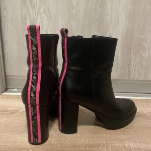 Svarta boots med rosa detaljer - Snygga svarta boots med hög klack och coola rosa detaljer. De har en unik ormskinnsmönstrad rem längs baksidan och sulan, vilket ger dem en edgy look. Perfekta för höst och vinter, och de har en dragkedja på sidan för enkel på- och avtagning. Passar både till vardags och fest!