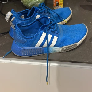 Blå Adidas NMD sneakers - Säljer ett par blå Adidas NMD sneakers i bra skick. De har den klassiska tre-randiga designen i vitt på sidorna och blå snörning. Skorna är bekväma och perfekta för vardagsbruk eller träning. Sulan är vit med blå detaljer och de har en modern look.
