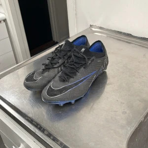 Svarta Nike mercurial fotbollsskor med blåa och silvriga detaljer - Säljer ett par svarta fotbollsskor med snygga blåa och silvriga detaljer. Skorna har snörning och är i bra skick. Perfekta för fotbollsträning eller match. Sulan har bra grepp för både gräs och konstgräs planer.