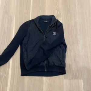 Svart tröja från J.Lindeberg - Säljer en snygg marin blå tröja från J.Lindeberg i storlek S. Tar gärna byten. Kan lösa ett bra pris vid snabb affär. Tröjan har en dragkedja framtill och är quiltad på framsidan. Perfekt för både höst och vinter, med en stilren design som passar till det mesta. Håll dig varm och trendig!