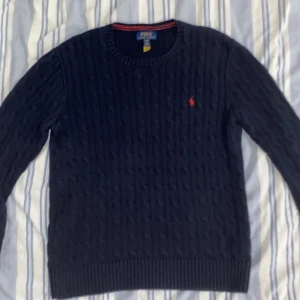Mörkblå stickad tröja från Ralph Lauren - Säljer en snygg mörkblå stickad tröja från Ralph Lauren. Den har ett klassiskt kabelstickat mönster och en broderad röd logga på bröstet. Perfekt för höst och vinter, med långa ärmar och ribbade kanter. Passar både till vardags och lite finare tillfällen. 🧶✨