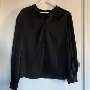 Svart blus med knappar. - Svart blus i skönt och luftigt material med knappar vid halsen och handlederna. Lite högre i kragen.