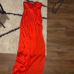 Röd axelbandslös långklänning - Säljer en stunning röd axelbandslös långklänning i satin. Perfekt för fest eller nyår! Den har en elegant slits på sidan och en tight passform som framhäver figuren. Klänningen är i mycket bra skick och redo att glänsa på nästa event.