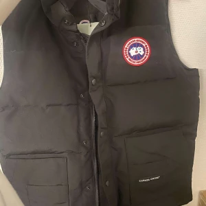 Svart dunväst från Canada Goose - Säljer en svart dunväst från Canada Goose i nytt skick. Västen har en klassisk design med dragkedja och knappar framtill. Perfekt för kyliga dagar och har den ikoniska loggan på bröstet. Kommer i originalförpackning.