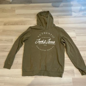 Olivgrön hoodie från Jack & Jones. Storlek 176  - Säljer en olivgrön hoodie från Jack & Jones i mycket bra skick. Den har en klassisk design med en stor logga på framsidan och en bekväm huva. Perfekt för höst och vår, och passar till både chill och casual outfits. Materialet känns mjukt och skönt mot huden.