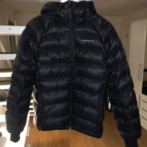 Svart dunjacka från Peak Performance - Säljer en snygg svart dunjacka från Peak Performance i mycket bra skick, i stort sett ny skick. Jackan har en dragkedja framtill och en praktisk huva. Perfekt för kalla vinterdagar och höstpromenader. Den är varm och bekväm, idealisk för både vardag och utomhusaktiviteter. Köpte för 2500kr. Pris kan diskuteras. Endast använt i nån vecka💕
