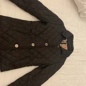 Svart quiltad jacka från Burberry Brit - Säljer en snygg svart quiltad jacka från Burberry Brit. Den har en klassisk design med knappar framtill och det ikoniska rutiga fodret. Perfekt för höst och vår! Jackan är i mycket bra skick och gjord av polyester och bomull. Vintage collection på burberry och 100% äkta 
