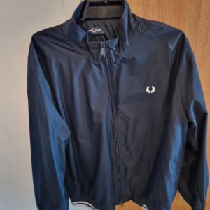 Mörkblå vindjacka från Fred Perry storlek xxl - Säljer en snygg mörkblå vindjacka från Fred Perry. Den har en dragkedja framtill och ett klassiskt broderat Fred Perry-märke på bröstet. Perfekt för vår och höst med sin lätta och stilrena design. Jackan har ribbade muddar vid ärmslut och nederkant för en skön passform.