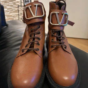 Bruna boots  - Säljer ett par snygga bruna boots från Valentino i skinn. De har en cool snörning och ett stort V-spänne i guld på framsidan. Perfekta för höst och vinter med sin robusta sula. Använd dem för att ge din outfit en lyxig touch!