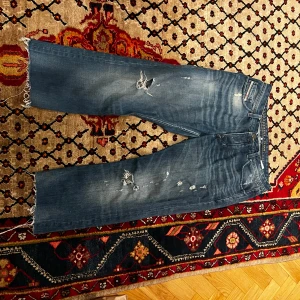 Blå jeans med slitningar - Säljer ett par blå jeans i storlek 38 med coola slitningar på framsidan. De har en rak passform och fransiga benslut för en avslappnad look. Perfekta för en casual stil och tillverkade i Italien. Passar bra till både vardag och fest!