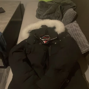 Moose Knuckles vit päls  - Säljer en svart dunjacka från Moose Knuckles med vit päls på huvan. Jackan har en cool broderad logga på insidan och snygga metallknappar. Perfekt för kalla vinterdagar! 🖤