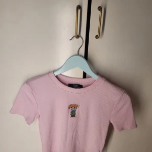 Rosa ribbad t-shirt från Bershka - Säljer en söt rosa ribbad t-shirt från Bershka med ett litet Rugrats-motiv på bröstet. Perfekt för en avslappnad stil. T-shirten är i mycket bra skick och har korta ärmar. Passar perfekt till vår och sommar!