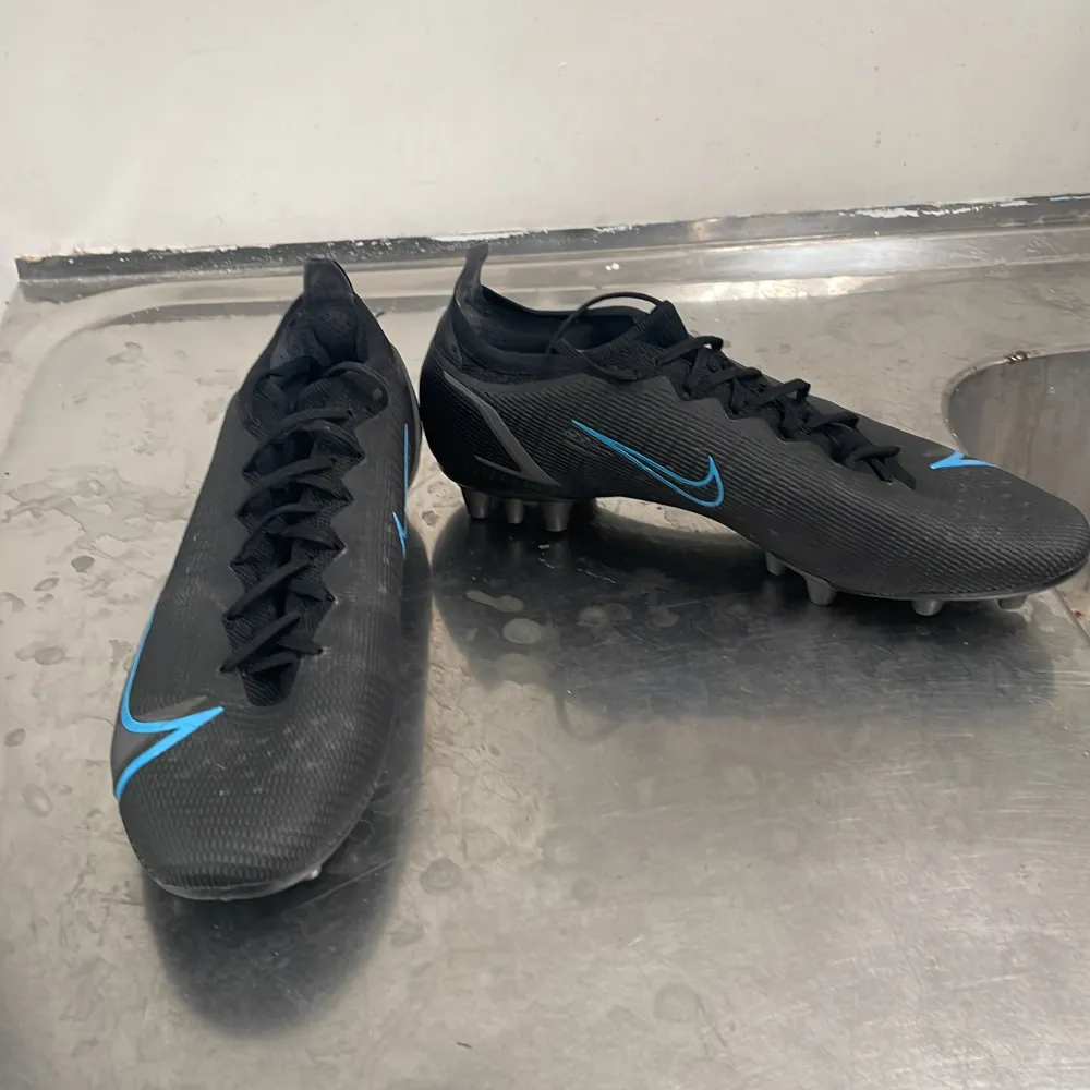 Säljer ett par svarta Nike fotbollsskor i syntetmaterial med snygga blå detaljer. Skorna har snörning och är i mycket bra skick. Perfekta för fotbollsträning eller match. De har en tight passform och är designade för att ge bra grepp på planen.. Skor.