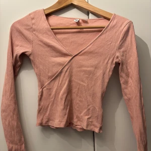 Rosa ribbad omlottopp från H&M - Säljer en superfin rosa ribbad omlottopp från H&M Divided. Den har en v-ringad design och långa ärmar, perfekt för både vardag och fest. Toppen är i bra skick och passar perfekt till vardags