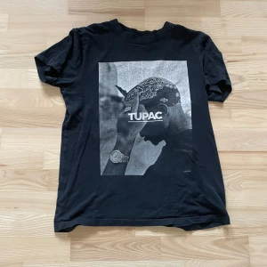 Svart T-shirt med Tupac-tryck - Säljer en svart T-shirt med ett stort tryck av Tupac på framsidan. T-shirten är kortärmad och i bra skick. Hör av dig vid frågor!