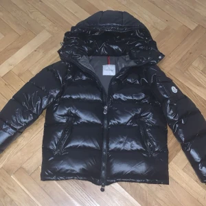 Svart pufferjacka från Moncler - Säljer en snygg svart pufferjacka från Moncler i mycket bra skick. Jackan har en glansig finish och är perfekt för kalla dagar. Den har en dragkedja framtill och en praktisk huva. Moncler-loggan syns tydligt på ärmen. Perfekt för vintern! ❄️