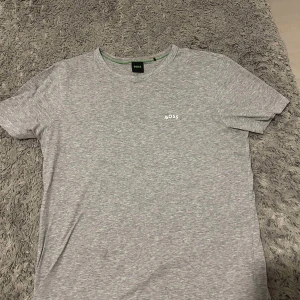 Grå t-shirt från Boss - Säljer en stilren grå t-shirt från Boss i mycket bra skick. Den är kortärmad och har en liten vit logga på bröstet. Perfekt för både vardag och avslappnade tillfällen. Passar bra till jeans eller shorts!