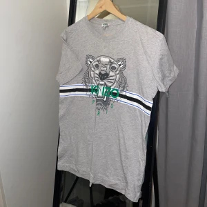Grå Kenzo T-shirt - Säljer nu denna snygga gråa Kenzo tishan med gröna och blåa nyanser i storlek S, funkar som unisex. Sparsamt använd, köptes för 1200kr, säljes nu för enbart 499kr. 🐯
