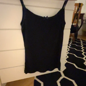 Svart ribbad linnetopp - Säljer en stilren svart ribbad linnetopp med smala axelband och en söt liten rosett framtill. Perfekt för sommardagar eller som ett lager under en kofta. Toppen är i bra skick och passar till många olika outfits!