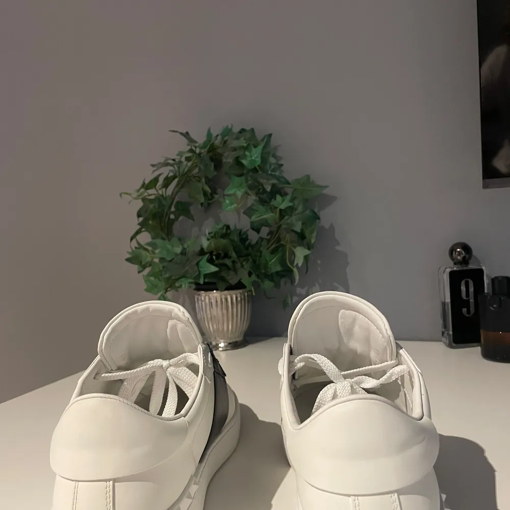 Säljer ett par snygga Valentino sneakers i vitt skinn med en bred svart rem över snörningen. Skorna har de ikoniska rockstud-detaljerna på hälen och är i mycket bra skick. Perfekta för både vardag och fest!. Skor.