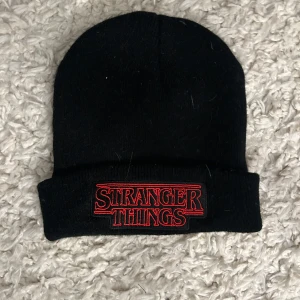 Svart Stranger Things mössa - Säljer en svart mössa med Stranger Things-logga i rött. Perfekt för fans av serien! Den är stickad och håller dig varm under kyliga dagar. Passar bra till både vardag och speciella tillfällen.